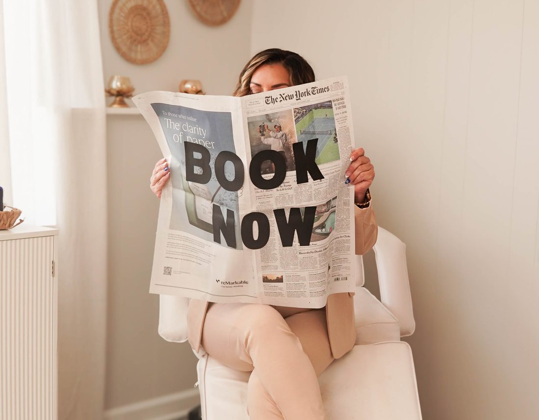 Christina holding a newspaper that reads Book Now in the Glamorous Grinz & Beauty studio